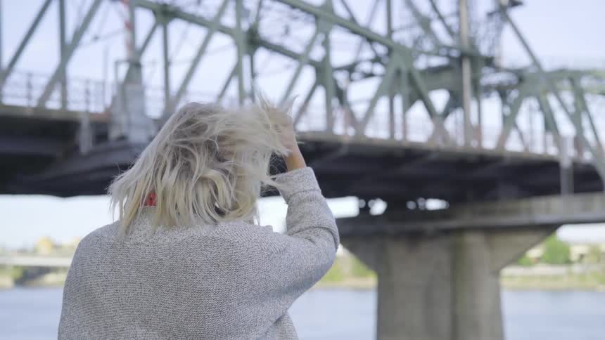 A young woman with shaggy blonde hair runs her fingers through it as she sits near a river and a steel and concrete bridge