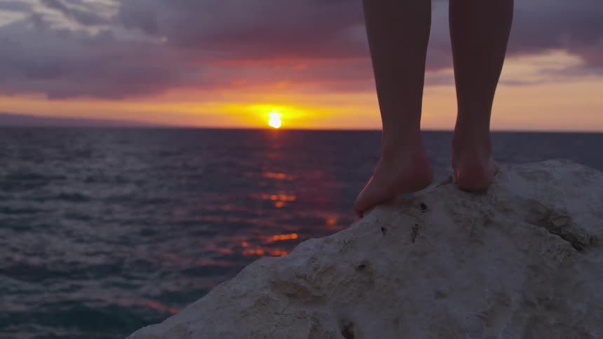 Person stands on a rock by the ocean with the sun setting