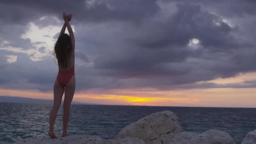 A young woman stands on a rock and stretches in front of the ocean