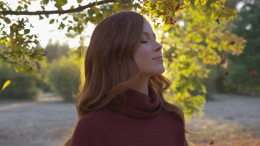 Woman stands with eyes closed as leaves fall around her