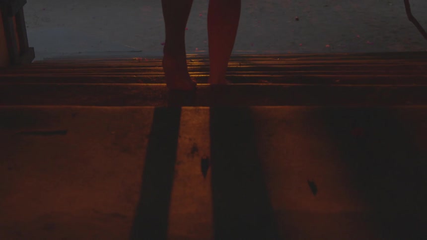 A barefoot woman walks slowly down a set of dirty stairs in the darkness while illuminated by only minimal light