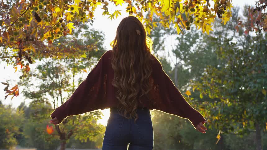 Woman raises her arms as leaves fall and the sun sets