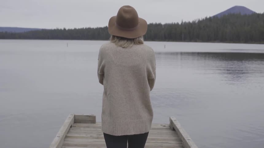 A woman with a sweater and a hat walking on a pier on a lake