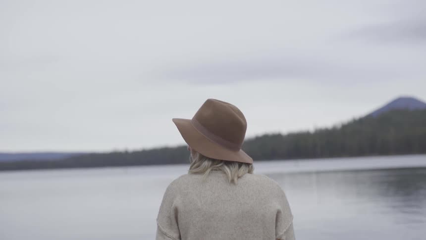 A blonde woman wearing a brown cat looks across a lake