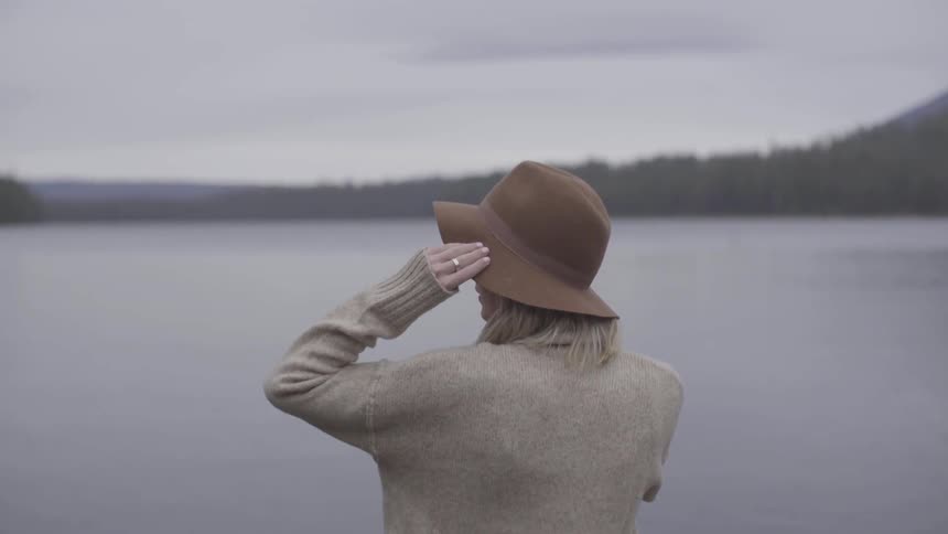 A woman holds onto her hat while staring at the mountains and lake