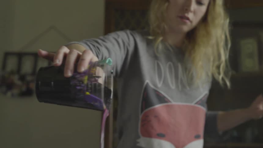 A blonde woman pours purple paint out of a cup