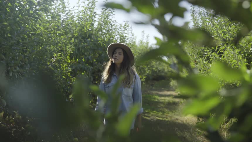 A brunette woman wearing a straw hat strolls down a path between green bushes