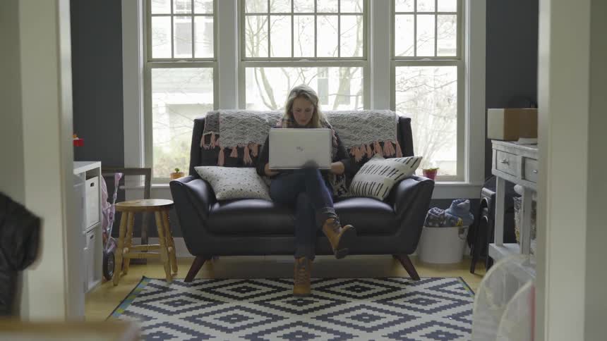 An attractive blonde woman walks to her couch and sits down and starts using her laptop computer