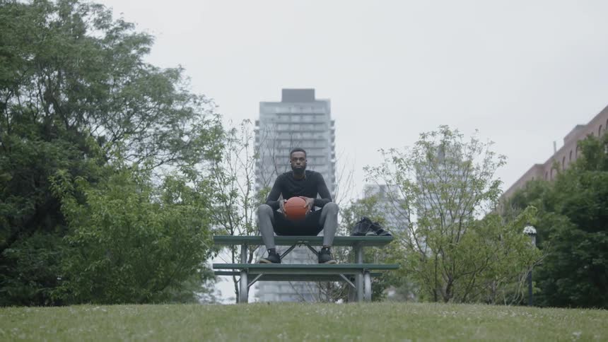 A young black man holding a basketball sits on a picnic table in a park