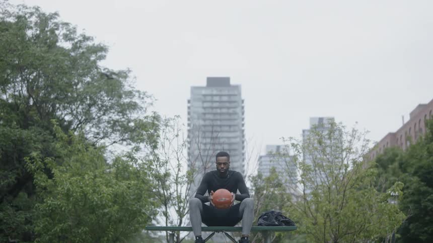 Black man sitting on a bench with holding a basketball near trees and buildings