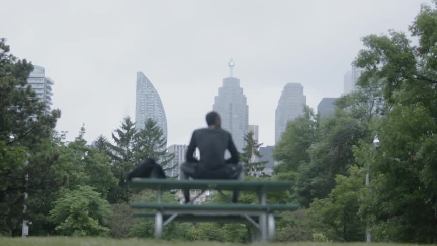 As he sits and ponders his life a black man sits atop a park bench with the city skyline peaking through the trees