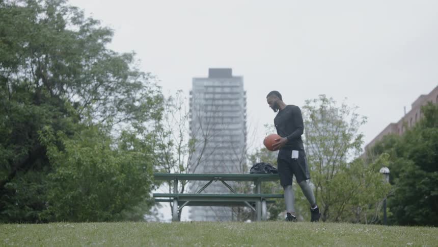 A black man holding a backpack and a basketball walks up and sits down on a picnic table