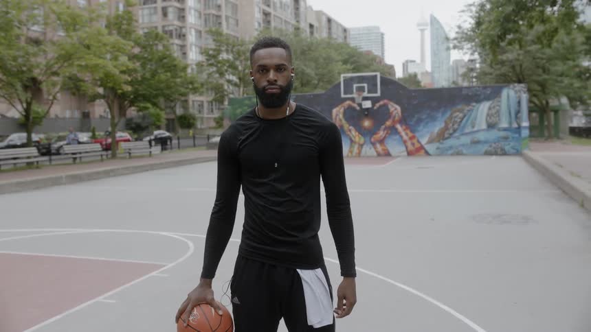 Bearded black man wearing earphones holds basketball and stands in the basketball field in the daytime