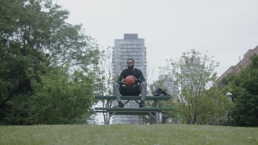 A man holding a basketball sits at a picnic table near a city skyline