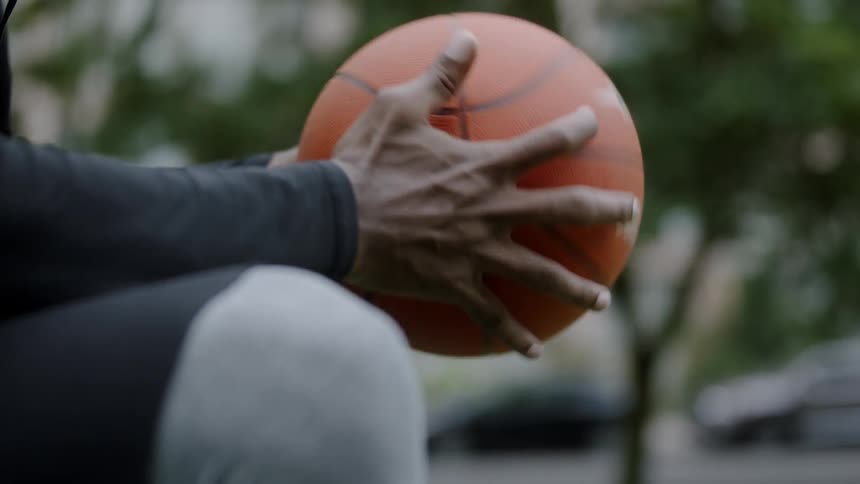 A black man sitting outdoors twirling a basketball in his hands