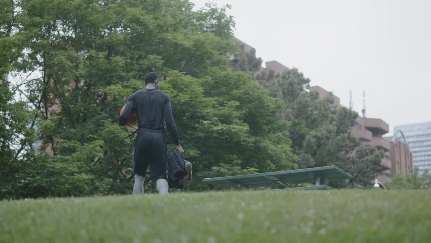 A black man holding a backpack and a basketball walks across park grass to a picnic table