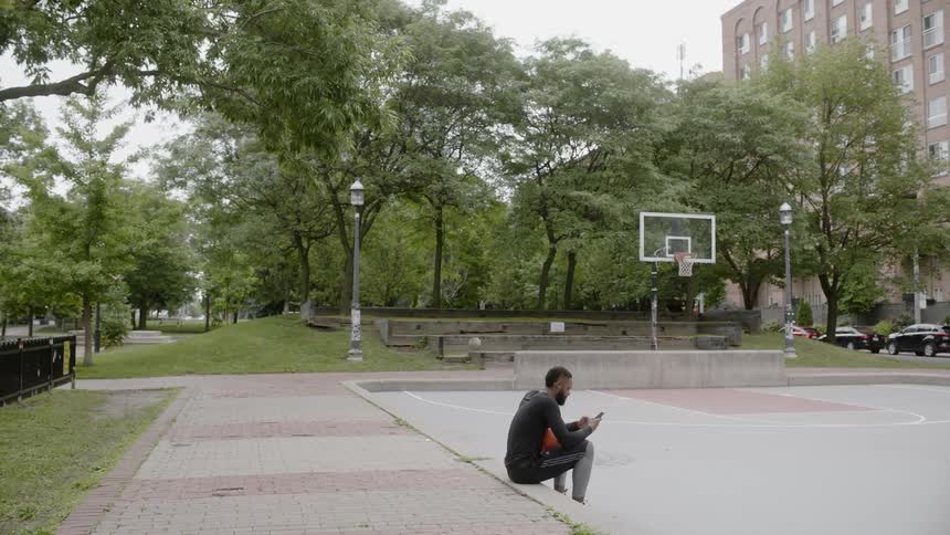 A black man sits on the side of a basketball court while using a smartphone