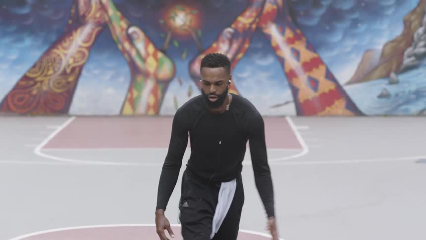 A black man dribbles and shoots a basketball on a basketball court with a mural painted on a wall behind him