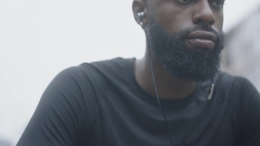 Bearded black man wearing earphones sits down and holds basketball outdoors in the daytime