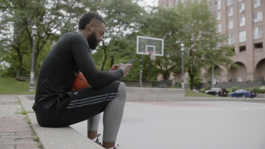Black man using his cellphone while sitting at a basketball court and holding a basketball on his lap