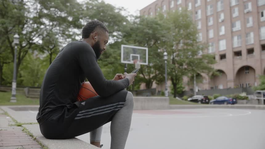 Bearded black man wearing earphones uses phone by the basketball field in the daytime