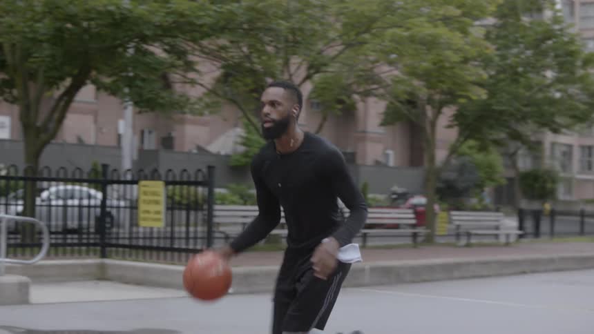 Bearded black man wearing earphones plays basketball in the basketball field in the daytime