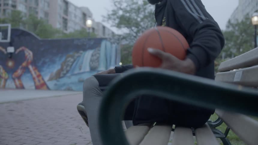 Bearded black man wearing earphones sits down and puts basketball on the bench at dusk