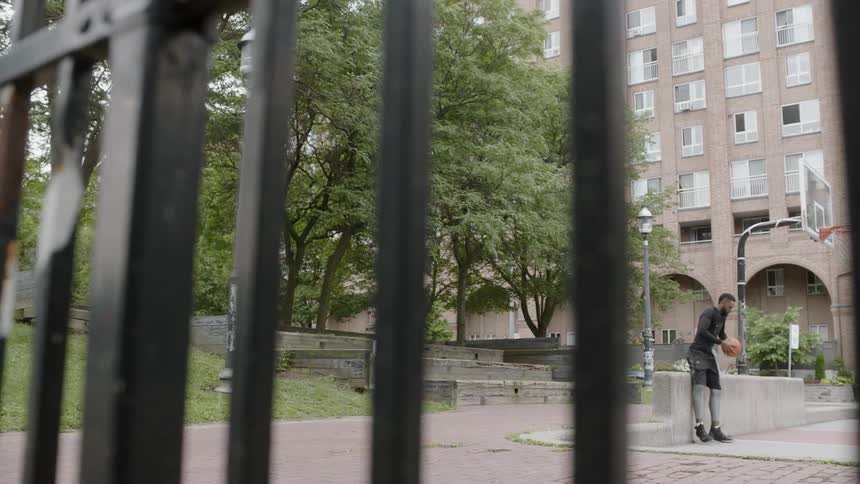 A black man sits on a concrete stoop bouncing a basketball and then stands up and walks across a basketball court while bouncing the ball