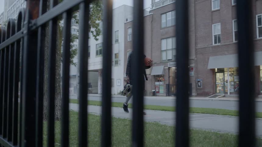 A man walks on the sidewalk carrying a basketball and extra shoes