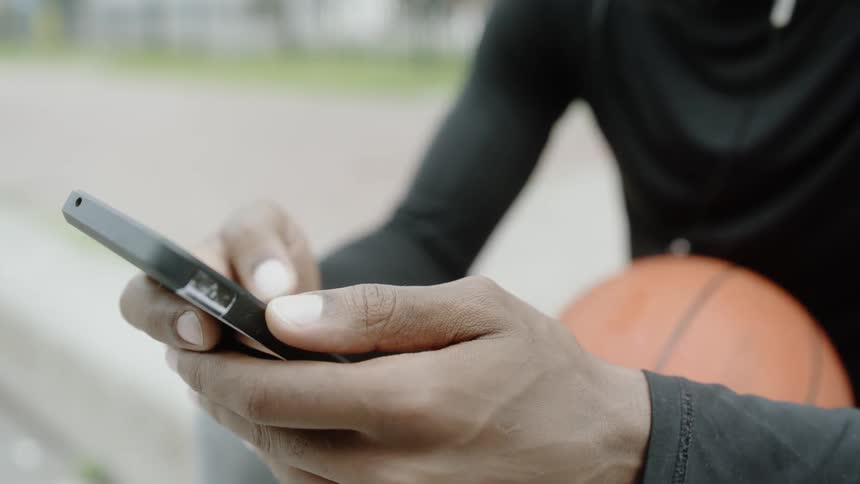 Black man using his cell phone while holding a basketball on his lap