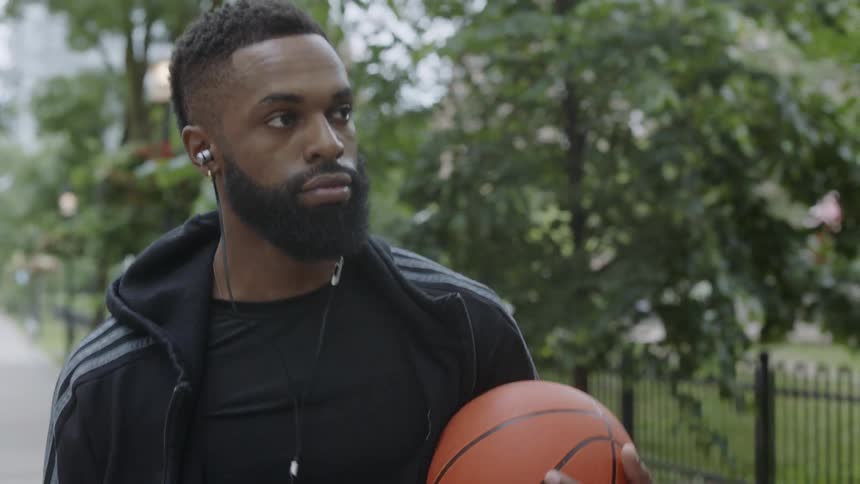 A black man walks through the city while carrying a basketball while wearing headphones