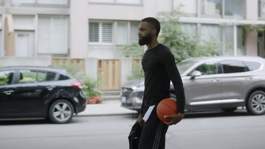 A man walks on the sidewalk of a neighborhood street carrying a basketball