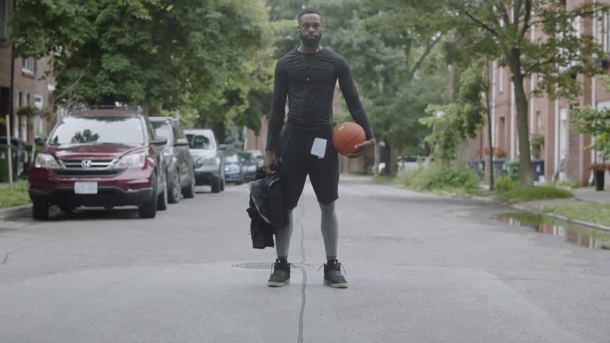 A black man stands in a street holding a basketball and a backpack
