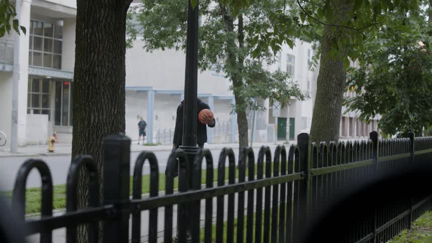 The man is walking on the sidewalk next to a black iron fence carrying a bag and his ball on a