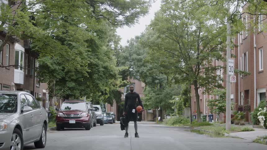 A man wearing black standing in the middle of a neighborhood street holding a basketball and a bag