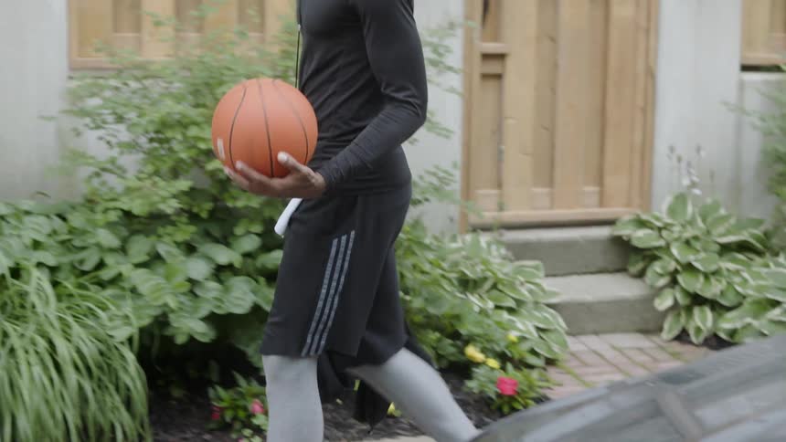 Bearded black man wearing earphones carries basketball and walks down the street in the daytime