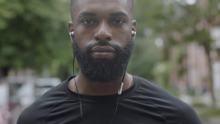 A young black man with ear buds in stands staring straight ahead