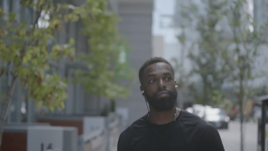 Bearded black man wearing earphones walks down the street in the daytime