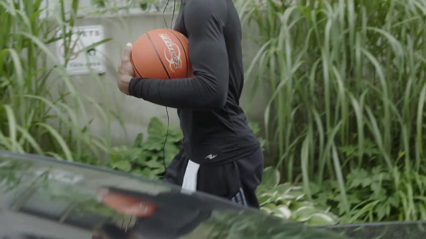 A young black man walks down a neighborhood sidewalk while carrying a basketball