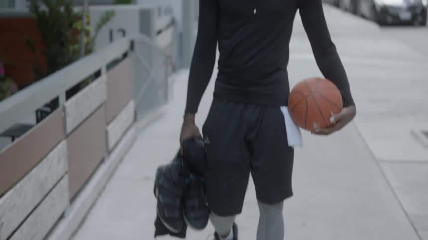 Bearded black man wearing earphones carries basketball and walks down the street in the daytime