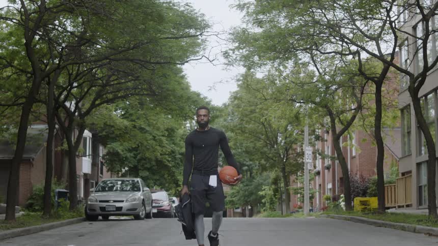 A man carrying a basketball and wearing headphones walks down a street