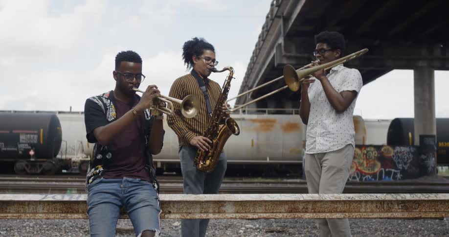 Three young black men play musical instruments by the railroad tracks