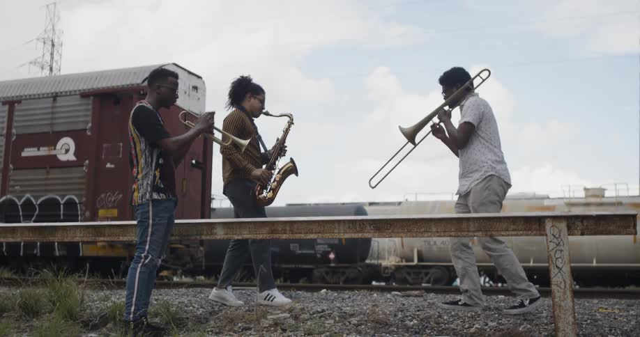 Three men play brass instruments in front of trains