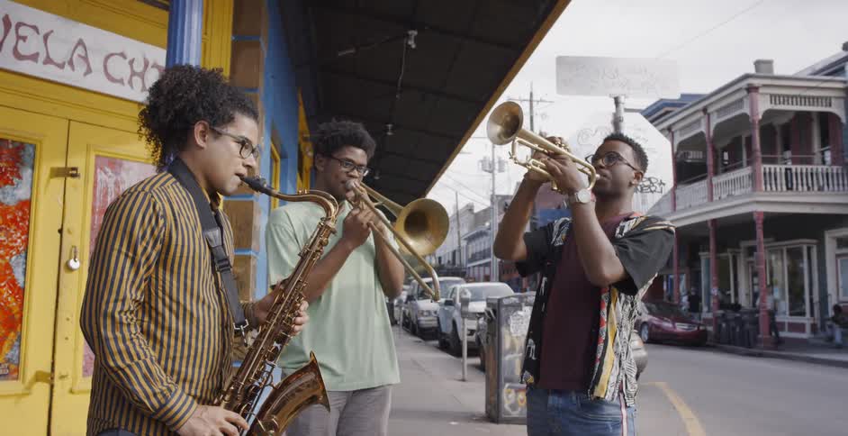 Three young black men stand on a street corner playing musical instruments
