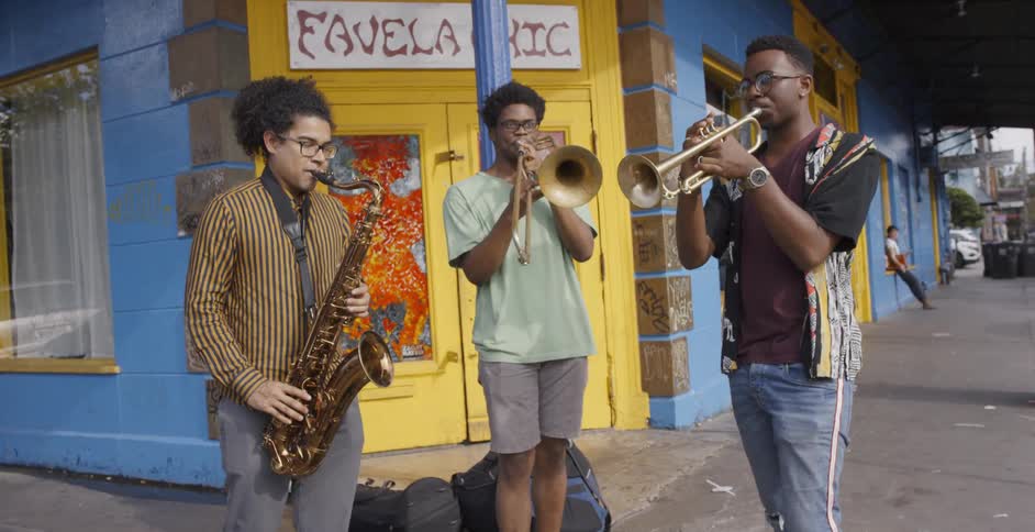 Three young black men stand on street corner playing musical instruments