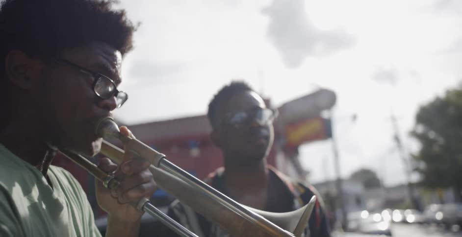 Two men one playing a trombone the other a trumpet stand on a sidewalk with buildings behind them