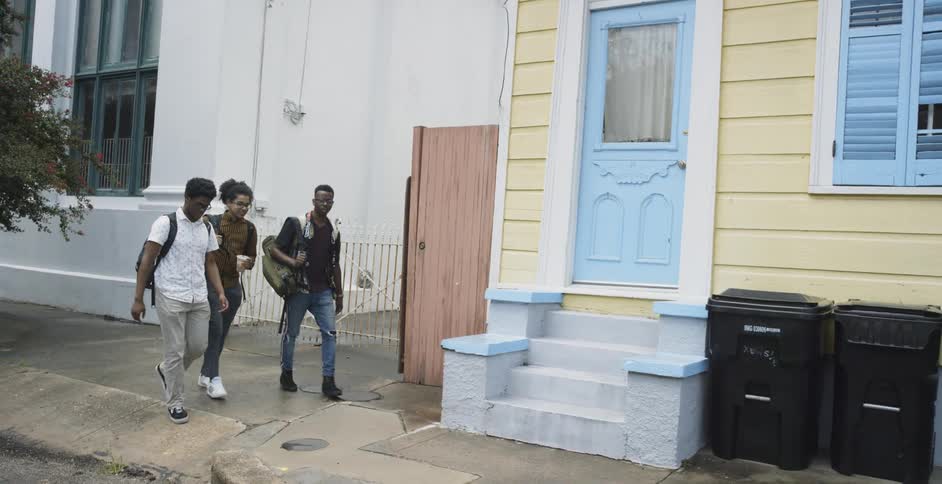 Three African American teenagers wearing backpacks walk down a sidewalk through a neighborhood