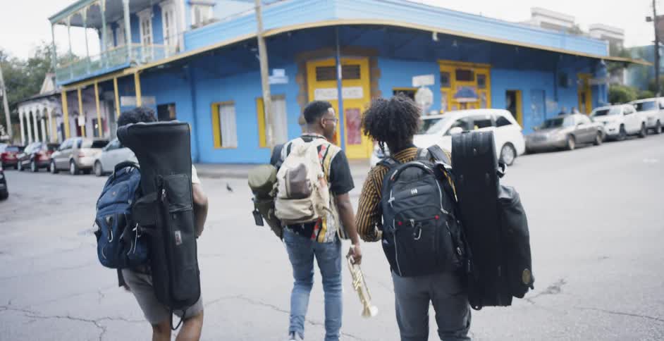 Three teenagers wearing backpacks and carrying musical instruments cross a street towards a blue building on the corner