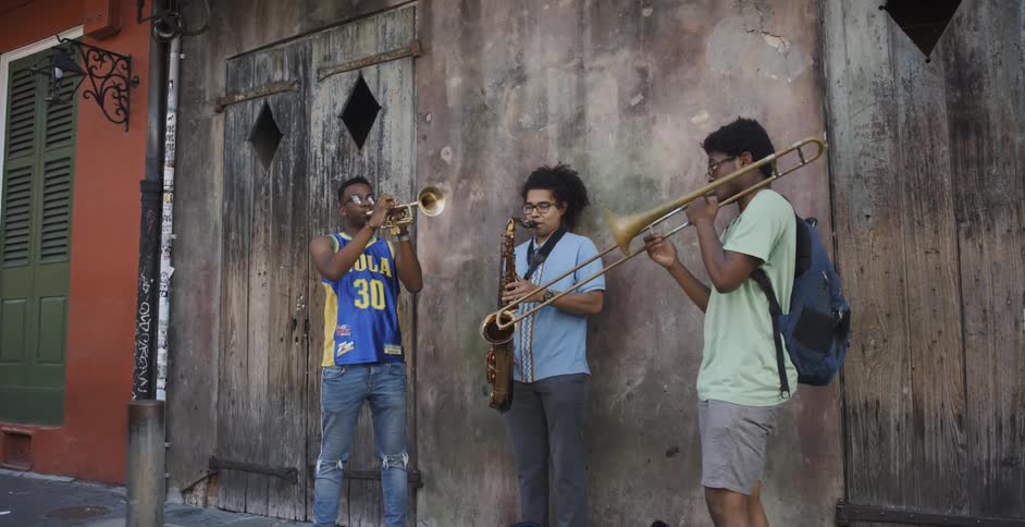 Three casually dressed young men stand along a street while playing the trumpet trombone and the saxophone