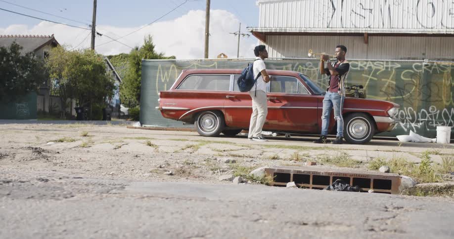 Two black men stand by a red car as one of them plays a trumpet